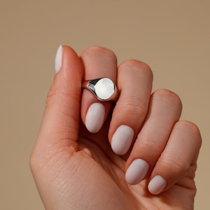 May include: A silver signet ring with a smooth, oval-shaped top. The ring is worn on a hand with light pink nail polish.