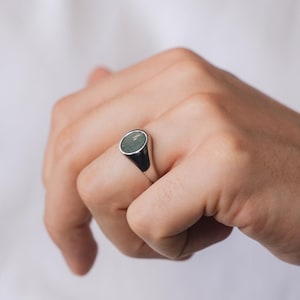 May include: A silver signet ring featuring an oval, green gemstone. The ring is worn on a finger. The ring has a polished finish and a classic design. The background is plain white.