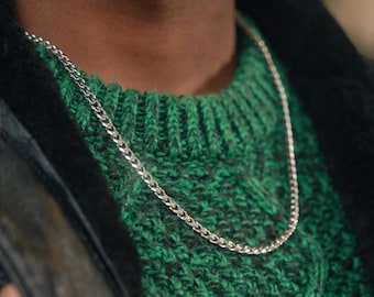 May include: A silver chain necklace with a curb link design. The necklace is worn over a green cable-knit sweater and is visible against a black jacket. The image is a close-up of the necklace and upper body.