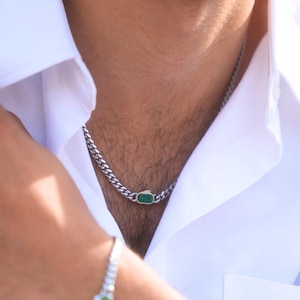 以下が含まれることがあります： シルバーチェーンのネックレスとブレスレットのセット。各ピースには、小さな楕円形のエメラルドグリーンの石が特徴です。ジュエリーは白い襟付きシャツの上に着用されています。チェーンのリンクはしっかりと組み合わされています。