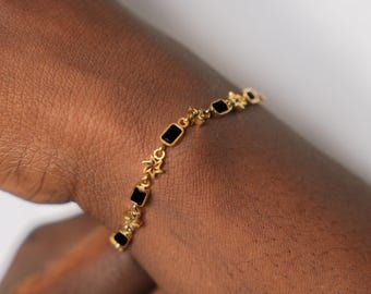 May include: Gold-toned bracelet featuring rectangular black stones and star-shaped accents. The bracelet is worn on a wrist, showcasing its delicate design and contrasting colors. A stylish piece of jewelry.