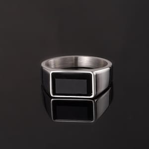 May include: A silver ring featuring a rectangular black stone set within a square bezel. The ring is presented on a reflective surface, highlighting its polished finish and geometric design. The ring's band is thick and robust.