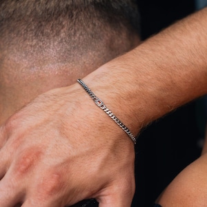 May include: A silver-toned curb chain bracelet featuring a small, rectangular link with a single embedded red stone. The bracelet is constructed from interlocking metal links that form a continuous band around the wrist, showcasing a metallic finish.