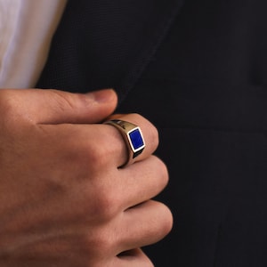 Puede incluir: Un anillo plateado con una piedra rectangular azul oscuro. El anillo se lleva en una mano, con una chaqueta oscura de traje en el fondo. El diseño del anillo es simple y elegante.