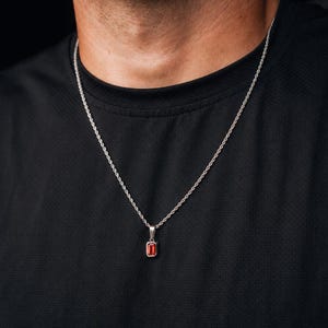 May include: A silver-toned rope chain necklace featuring a small, rectangular pendant with a vibrant red gemstone set in a simple metal frame. The necklace rests against a black sleeveless shirt, highlighting the contrast between the metal chain and the bright red stone.