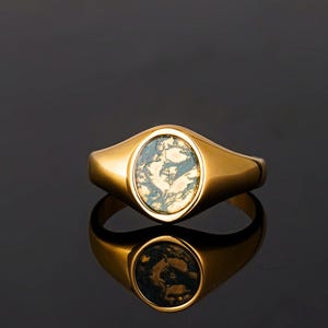 May include: A gold signet ring with an oval moss agate stone. The stone has green and white inclusions. The ring is shown on a reflective black surface.