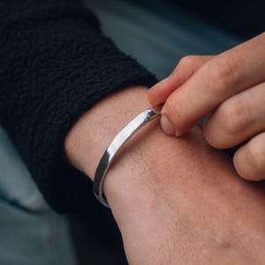 May include: A polished silver cuff bracelet and a matching ring. The bracelet is a simple, curved band. The ring is a thin band. The person is wearing a black jumper.