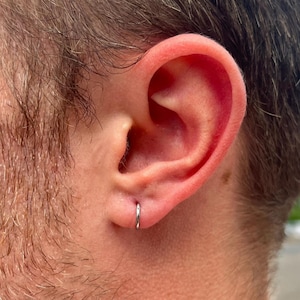 May include: A close-up of a person's ear with a small, silver hoop earring. The earring is a simple, thin band. The ear is pinkish-toned, and the person has short, dark hair and a beard.
