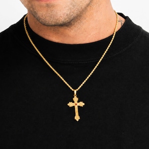 May include: A gold-toned rope chain necklace featuring an ornate cross pendant. The cross has intricate decorative detailing along its edges and a small, circular red stone set at the centre where the two bars of the cross intersect.