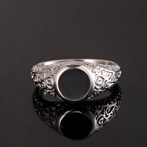 May include: Silver ring with a black, oval centre stone. The ring has detailed, swirling patterns around the band. The ring is shown on a reflective surface, creating a mirrored effect.