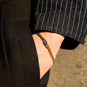 Puede incluir: Pulsera de cadena dorada con una piedra rectangular de color turquesa. La pulsera se lleva en la muñeca, con un traje negro y una chaqueta de rayas finas en el fondo. Es una joya.
