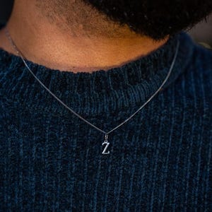 May include: A silver necklace with a small pendant in the shape of the letter "Z" hangs against a dark blue sweater. The chain is delicate, and the pendant is centered. The background is blurred, focusing on the necklace.
