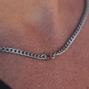 May include: Silver chain necklace featuring a small, square, red gemstone pendant. The necklace has a curb chain design and is worn around the neck. The gemstone is set in a square silver frame.