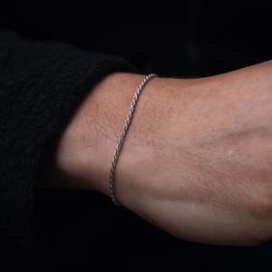 Könnte beinhalten: Ein silberfarbenes Seilarmband wird am Handgelenk getragen. Das Armband besteht aus eng verdrehten Metallsträngen. Der Hintergrund ist dunkel und betont die helle Farbe und Textur des Armbands. Das Armband ist ein schlichtes, elegantes Schmuckstück.