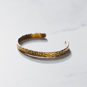Pode incluir: Uma pulseira de punho dourada com uma superfície texturizada. A pulseira aberta está colocada sobre um fundo de mármore branco. A pulseira tem um design martelado.