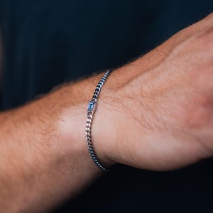 May include: A silver-toned curb chain bracelet featuring a single rectangular light blue gemstone set in a metallic frame. The bracelet consists of small, uniform interlocking links that wrap around the wrist, creating a sleek and simple design.