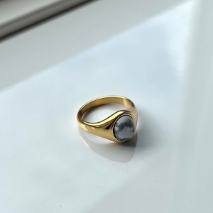 Op de afbeelding: Een gouden ring met een witte steen in het midden. De ring is simpel en elegant.