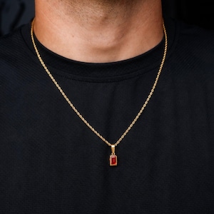 May include: A gold rope chain necklace featuring a rectangular pendant with a deep red gemstone set in a gold frame. The necklace rests against a plain black shirt, highlighting the contrast between the metallic gold finish and the red stone.