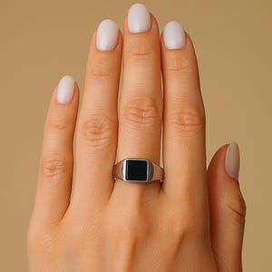 Könnte beinhalten: Ein silberner Siegelring mit einem schwarzen quadratischen Stein.