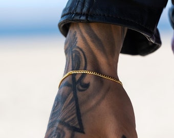 May include: A gold chain bracelet on a person's wrist with a tattoo of an ankh and a triangle. The person's hand is also tattooed with a cross.