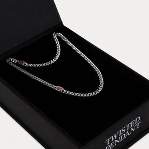 Puede incluir: Un collar de cadena de metal plateado con dos eslabones rectangulares con piedras rojas incrustadas, presentado dentro de una caja negra con el texto TWISTED PENDANT impreso en blanco en el lateral de la tapa.