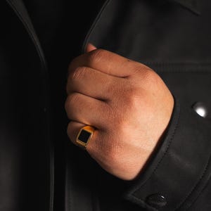 Könnte beinhalten: Ein goldener Siegelring mit einem schwarzen quadratischen Stein.