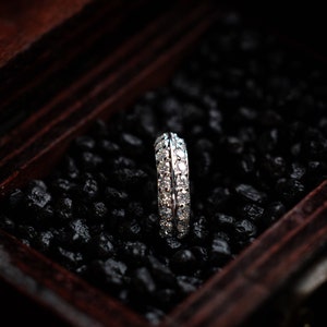 Peut inclure: Une bague en argent avec une double rangée de petits diamants clairs sertis dans une bande de métal lisse. La bague est présentée dans une boîte en bois foncé avec des cailloux noirs.