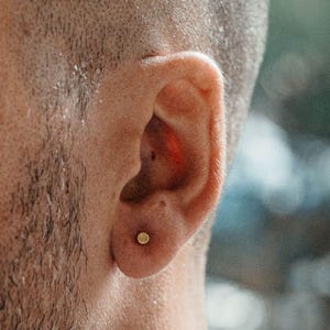 May include: A small, circular, gold-toned stud earring is centered on the earlobe. The earring features a smooth, flat, polished surface that reflects light, creating a subtle metallic sheen against the skin of the ear.