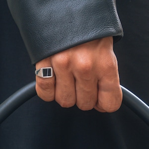 以下が含まれることがあります： 黒い正方形のセンターが付いたシルバーリングが指に着用されています。リングは長方形で、平らな上部があります。その人は黒い革のジャケットを着ています。