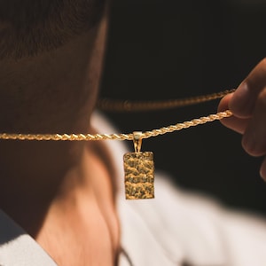 May include: A gold-colored necklace with a textured rectangular pendant. The chain is a twisted rope design. The pendant is held by a person wearing a light-colored shirt.