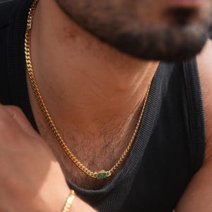 以下が含まれることがあります： 小さな長方形の緑色の石のペンダントが付いた、金色のチェーンネックレス。 ネックレスは黒いタンクトップの上に着用されています。 チェーンはカーブリンクのデザインです。 画像はネックレスと上半身のクローズアップです。