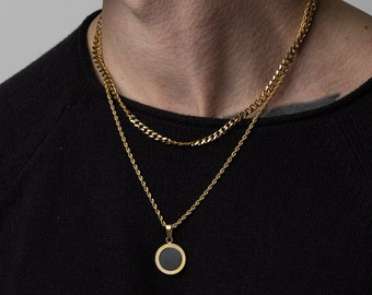 May include: Gold-toned necklace set. The top chain is a curb link style, and the bottom chain is a rope style with a circular pendant. The pendant has a black center and a textured gold border. The person is wearing a black sweater.