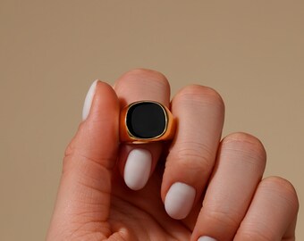 May include: A gold-colored ring with a square, black stone. The ring is held up against a neutral background. The ring is a classic signet style.