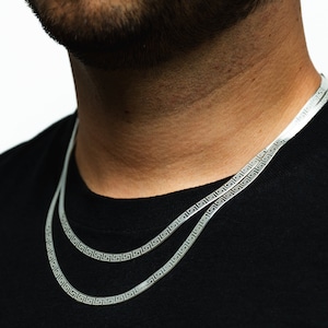 Puede incluir: Dos collares plateados con un patrón de llave griega se muestran sobre una camiseta negra. Los collares son planos y tienen un diseño en espiga. Los collares se llevan alrededor del cuello.