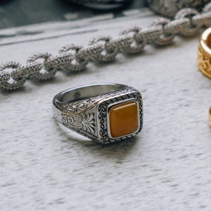 Puede incluir: Un anillo de plata con un diseño adornado y una piedra preciosa cuadrada amarilla. El anillo está sobre una superficie blanca con otros anillos de plata y oro.