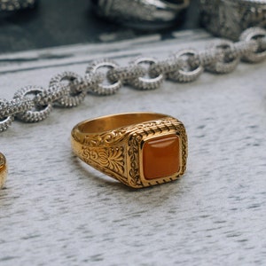May include: A gold ring with an orange gemstone set in a square bezel. The ring has intricate floral designs around the stone.