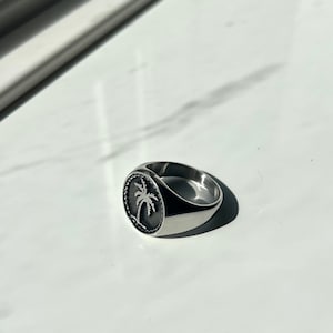 Könnte beinhalten: Ein silberner Siegelring mit einem Palmenbaum-Design. Der Ring ist oval geformt und hat eine glatte, polierte Oberfläche.