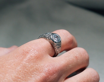 May include: A silver ring with an ornate design and a clear gemstone. The ring is on a person's finger.