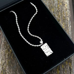 Puede incluir: Un collar plateado con un colgante rectangular texturizado descansa dentro de una caja de joyería negra. La cadena tiene un diseño de cuerda trenzada. El colgante tiene una apariencia de metal martillado, reflejando la luz.