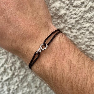 May include: A black cord bracelet with a silver clasp. The bracelet is worn on a person's wrist.
