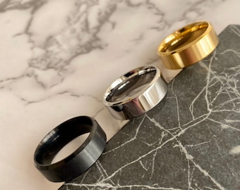 Peut inclure: Trois bagues de couleurs différentes : noire, argentée et dorée. Les bagues sont présentées sur une surface sombre, semblable à du marbre gris, avec un fond en marbre blanc et gris.