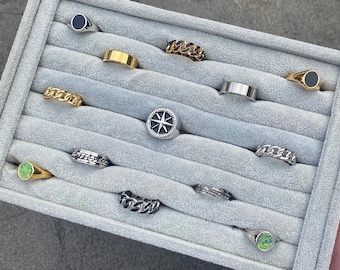 May include: A variety of rings displayed in a gray velvet tray. The rings include gold, silver, and black designs, with some featuring chain links, black onyx, and a compass rose. The rings are in various sizes and styles.