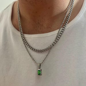 Può includere: Un set di collane argentate. La catena superiore è in stile a maglia a catena, e la catena inferiore ha un ciondolo con una pietra preziosa rettangolare verde smeraldo. La persona indossa una canotta bianca.