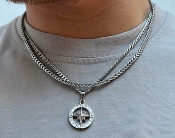May include: A silver necklace set comprising a chain and a compass pendant. The pendant features a star design and directional markings. The necklace is worn over a grey t-shirt.