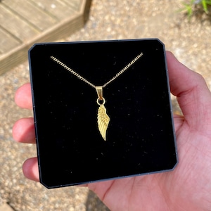 Könnte beinhalten: Eine goldene Kettenkette mit einem goldenen Flügelanhänger. Die Kette wird in einer schwarzen Samtbox präsentiert.