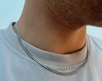 Op de afbeelding: Een zilveren kettingketting die om de nek van een persoon wordt gedragen. De ketting is van het curb-type met een platte, rechthoekige schakel.