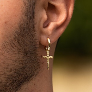 May include: A gold hoop earring with a dangling cross charm. The cross is detailed with a textured design.