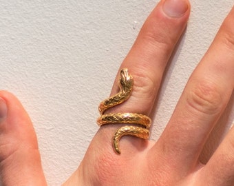 Puede incluir: Un anillo de serpiente de color dorado con una superficie texturizada. El anillo se lleva en el dedo índice de una mano.