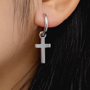 May include: Silver hoop earring with a dangling cross charm. The earring is worn in the left ear.