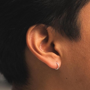 Puede incluir: Primer plano de una oreja con un pequeño pendiente de aro plateado. El pendiente tiene un diseño simple y circular. La persona tiene el pelo y la piel oscuros.
